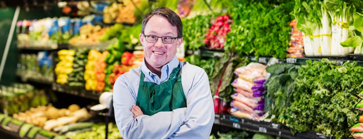 Man, in uniform with arms crossed, in fresh food section of supermarket.