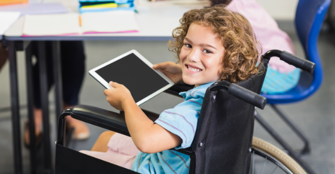 A boy sits in his wheelchair holding an ipad while smiling at the camera.