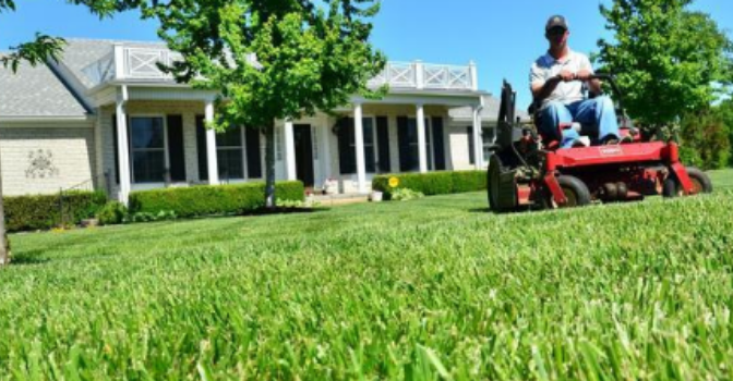 A man on a ride on mower is mowing a lawn in front of a house on a sunny and clear day.