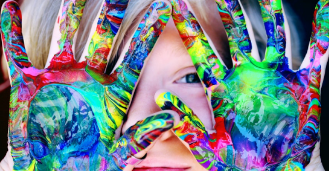 A child has his hand in front of his face which are covered in bright vibrant paint.