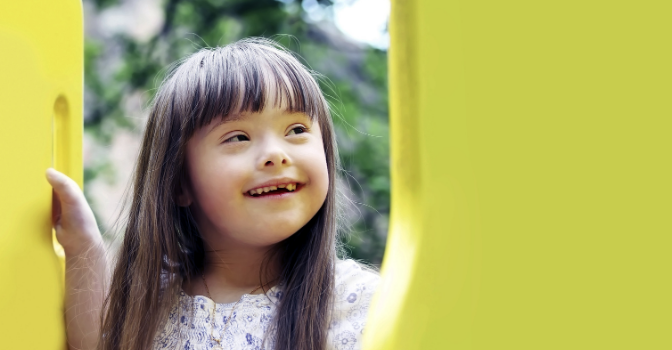 A young girl with long brown hair and Down syndrome is smiling and looking off camera while standing on a yellow playset in a park.