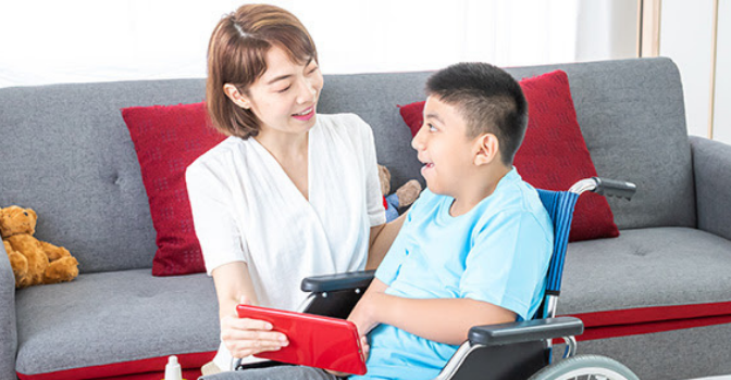 A boy in a wheelchair is looking up and smiling at a woman holding a tablet in front of them both.
