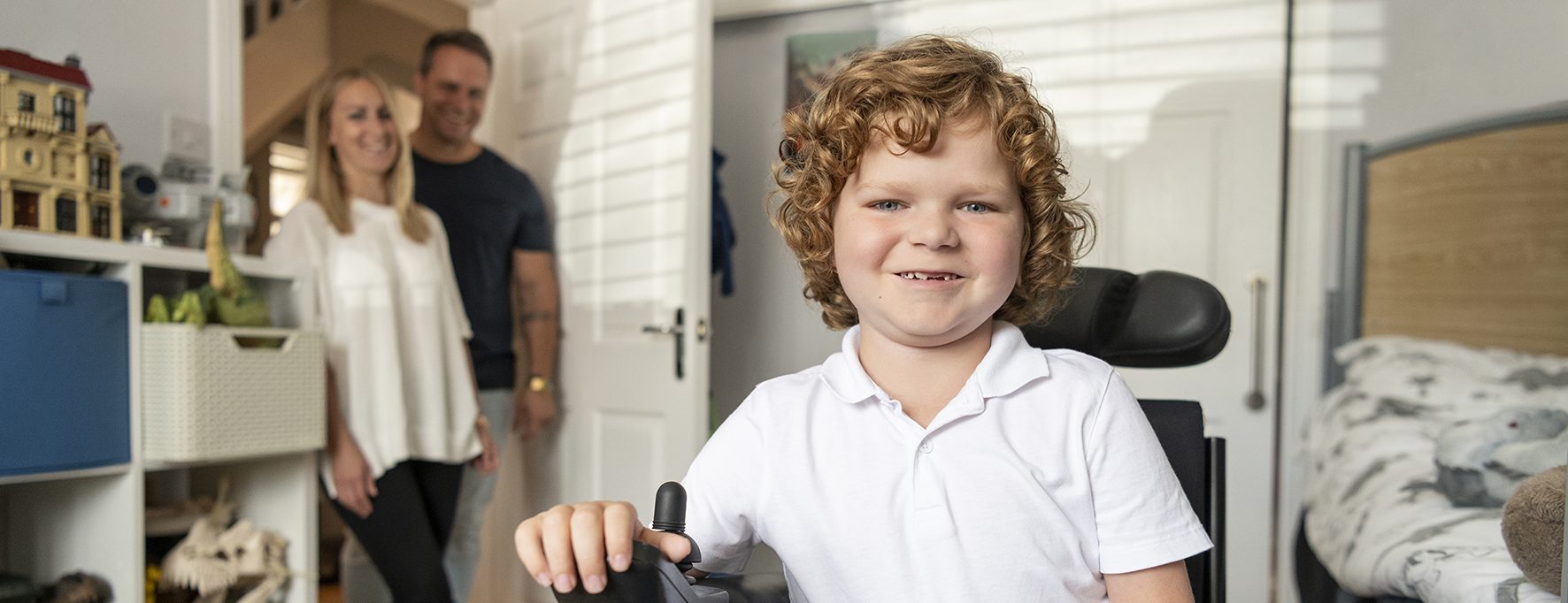 Cheerful young boy with muscular dystrophy in his bedroom, using powered wheelchair, mother and father smiling and watching in the background