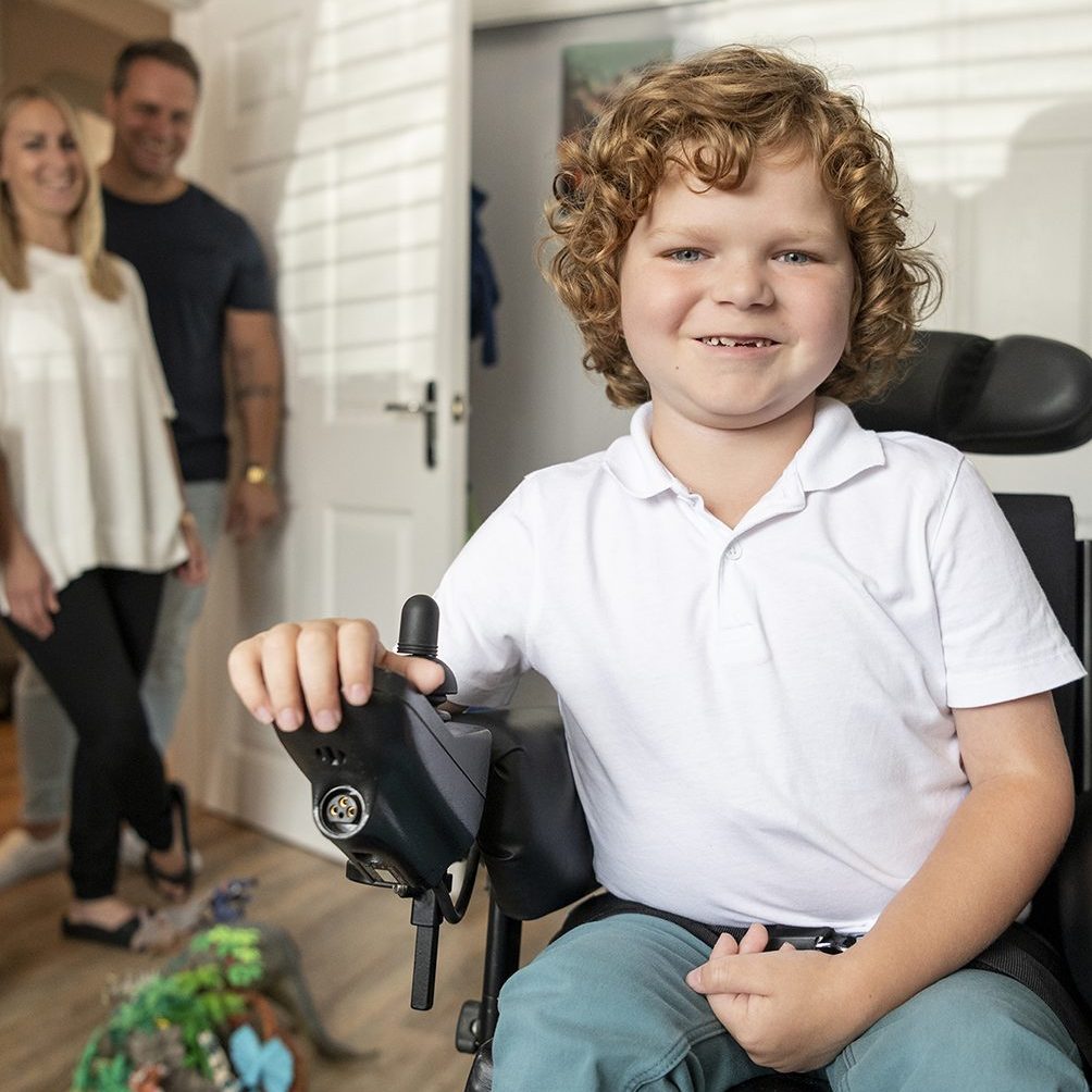 Cheerful young boy with muscular dystrophy in his bedroom, using powered wheelchair, mother and father smiling and watching in the background