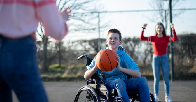 A boy in a wheelchair is playing basketball with 2 friends and it about to pass the ball to a person that is only half in the frame.