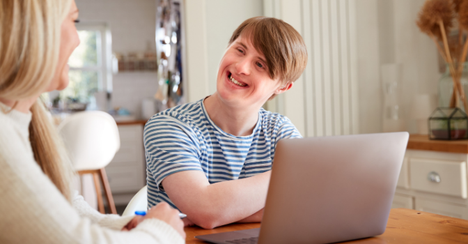 A man in a blue and white striped shirt is sitting in front of a laptop while smiling to someone off camera.