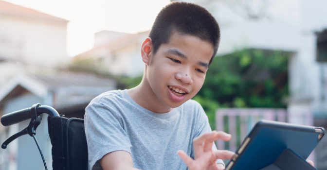 A boy sitting in his wheelchair outside in the day time is using a tablet and smiling.