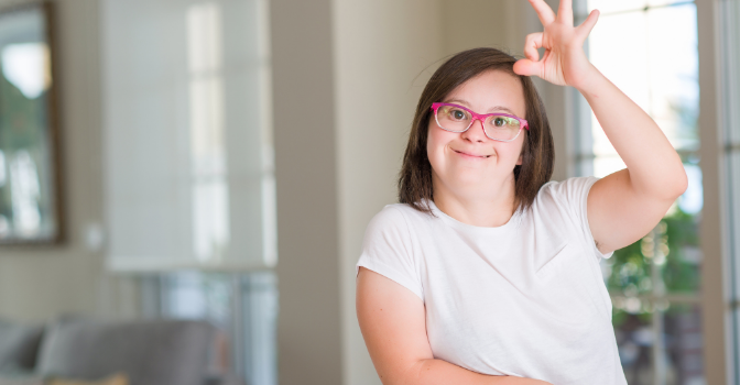 A girl wearing pink glasses and a white shirt is smiling at the camera giving the hand signal for OK.