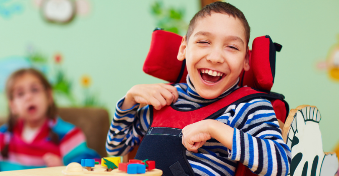 A boy sits in a red wheelchair while smiling toward the camera. He has a tray of toy blocks in front of him.