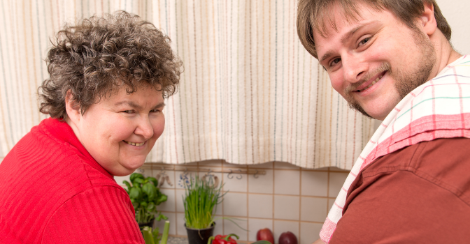 A woman and her carer are smiling at a camera while standing in a kitchen.