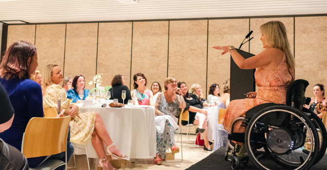 Karni Lidell is on stage in her wheelchair in a coral toned dress presenting to a room of mama's sitting at round dining tables with white tablecloths in The Calile Hotel ballroom.