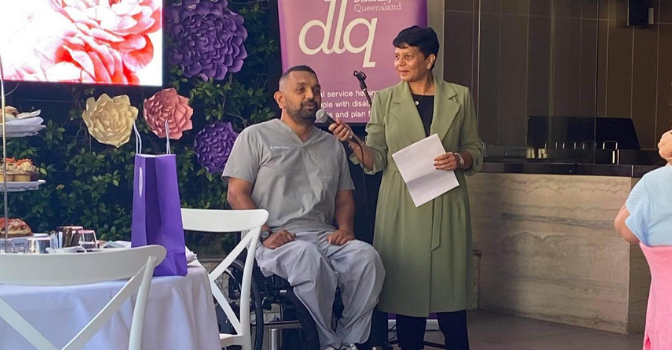 Dr Dinesh Palipana & his mum Chithrani Palipana speaking at the DLQ event. Dr Dinesh is dressed in grey scrubs and Chithrani is dressed in a light green dress. They are in front of the event banner with a garden wall in the background.