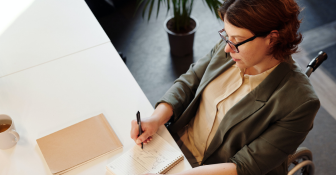 A lady with glasses in a wheelchaiar sits at a desk working on some paperwork.