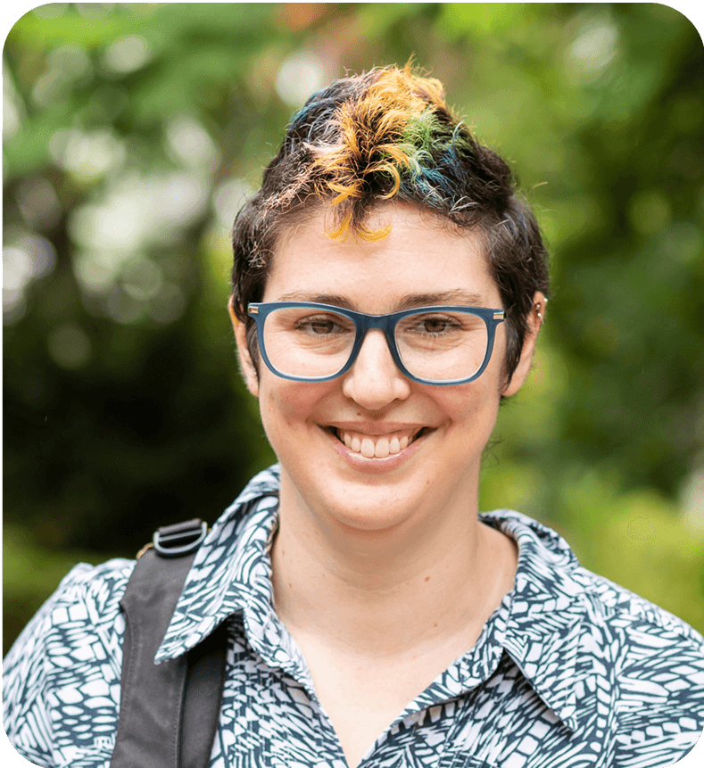 A young woman with some colour in her hair, standing outside with a backpack slung on her shoulder