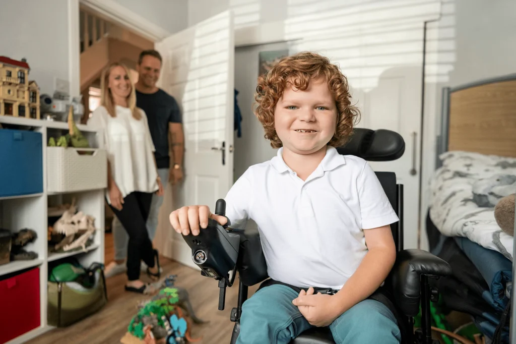 A young boy in an electric wheelchair in his bedroom, smiling with his parent in the doorway, smiling