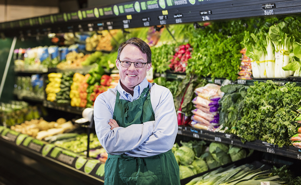 Man in an apron in front of the fruit and vegetable section in a supermarket