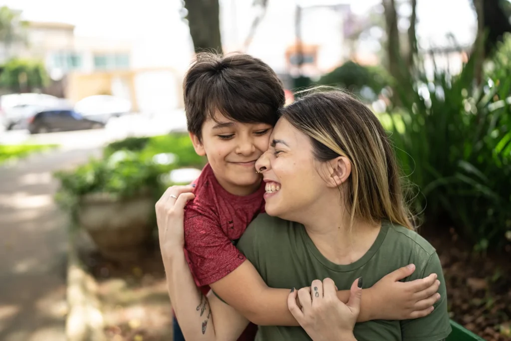 Mother and son smile and hug each other while sitting in a green leafy park.