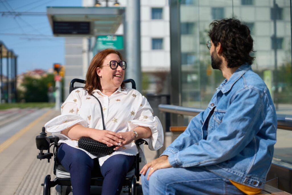 Woman in wheelchair chatting with friend at tram stop.
