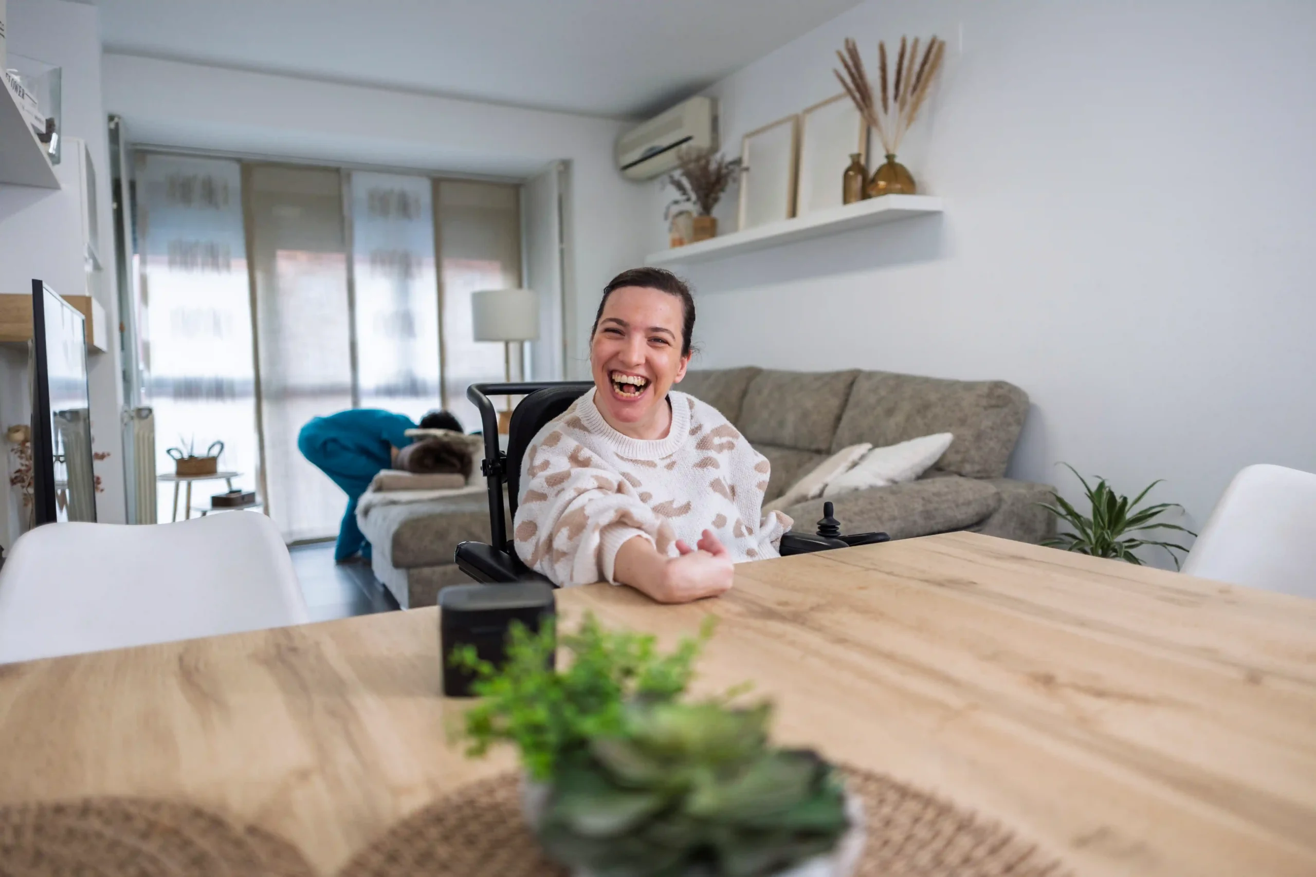 Young woman using a powered wheelchair smiling at a dining table in a bright, modern home.