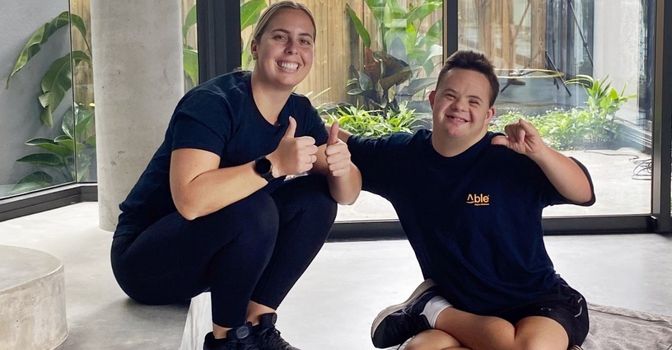 An Able Digital Wellness participant smiles and forms a hang ten symbol with his hand while his allied health professional gives two thumbs up and smiles next to him.