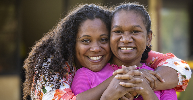 Two young aboriginal women outdoors with their arms around each other smiling at the camera.