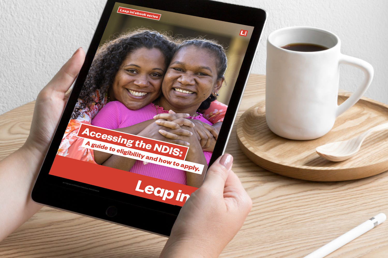 A black iPad with the Leap in! Accessing the NDIS ebook cover behing held on a table with a cup of coffee also sitting on the table.