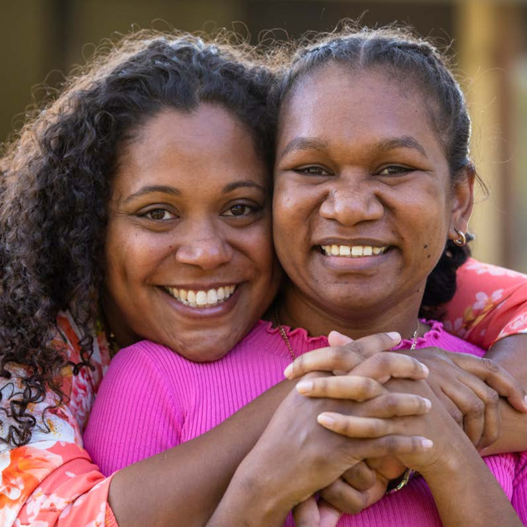 Two young aboriginal women outdoors with their arms around each other smiling at the camera.