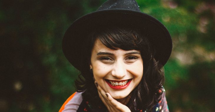 A woman with black hair and a black felt hat smiles and rests her hand on her face.