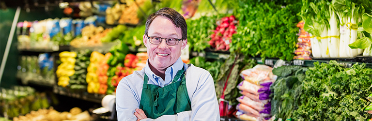 Man, in uniform with arms crossed, in fresh food section of supermarket.