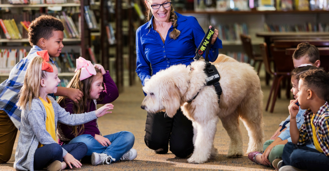 Teacher holding a therapy dog surround by children sitting on the ground.