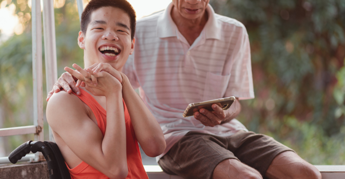 Young person with disability, on balcony outside, with parent showing them a phone.