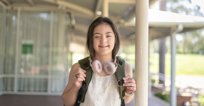 Young girl with disability standing outside with headphones and backpack on.