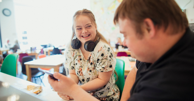 Young girl with disability wearing head phones, looking a a friend holding a phone.