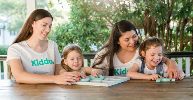 Two adults wearing Kiddo t-shirts, playing at a table with two young children.