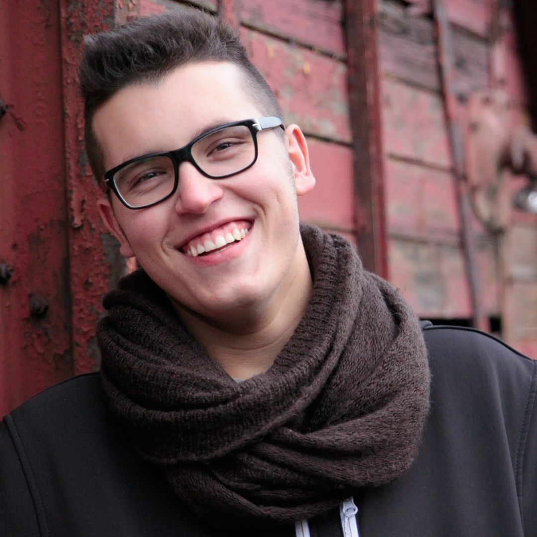 Young adult wearing a brown scarf and glasses, leaning against a wall and smiling