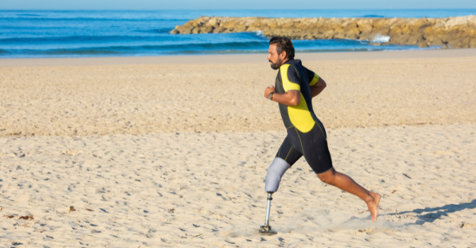 A man with a prosthetic limb runs along the beachfront in a wetsuit on a clear blue day.