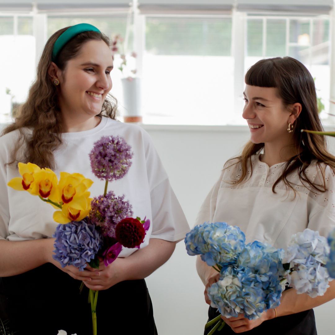 Two women smiling at each other arranging brightly coloured flowers