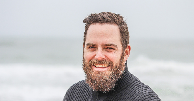 A man with a beard is smiling with the ocean in the background.