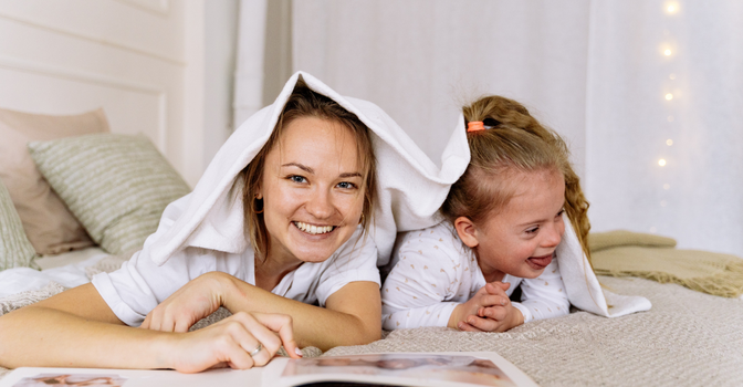 A mother and daughter are laying down laughing with a book and a blanket.