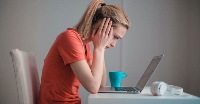 A woman sits at her laptop with her head in her hands, looking worried.