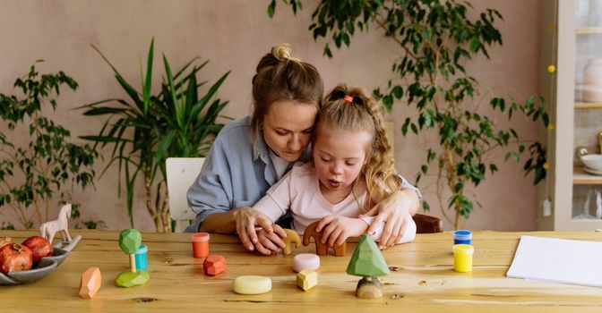 A mum and her daughter are playing happily with wooden sensory toys at a dining room table. There are plants in the background and natural light coming in from the right hand side.