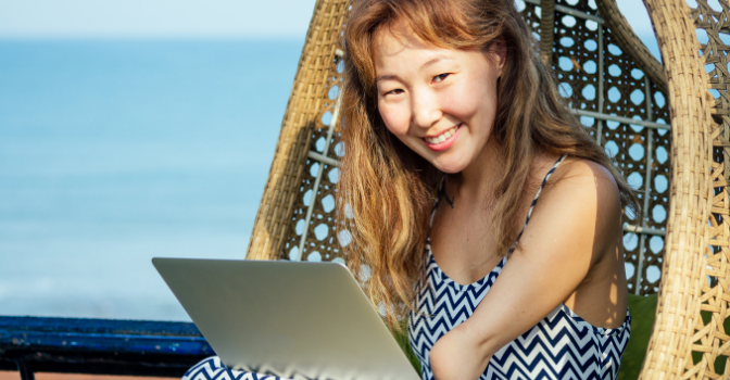 A lady with a partial limb sits on a rattan egg chair while on her laptop smiling at the camera.