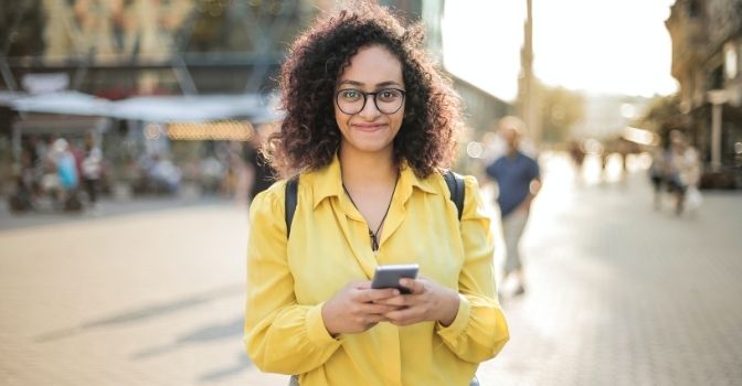 An image of a woman in a yellow shirt holding her phone and smiling.