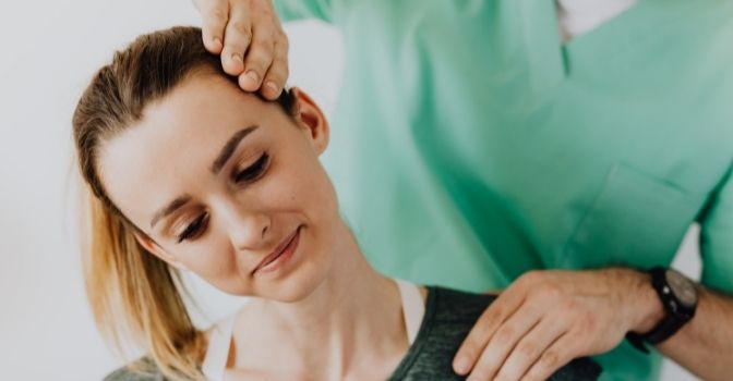 A woman is smiling with her head to the side as an allied health professional does work on her neck.