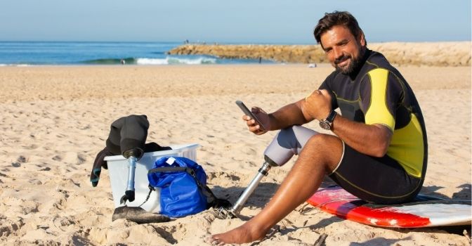 A man sits on the beach with his phone in his hand and is facing the camera and smiling.