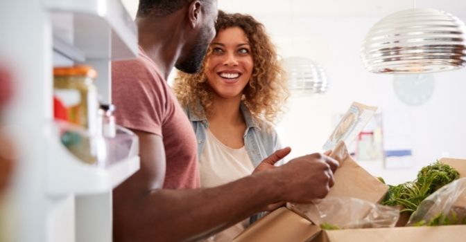 A man and a woman are in a kitchen unpacking a box of ready made food. The woman is looking at the man but facing the camera smiling happily.