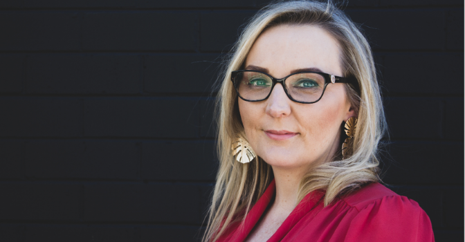 Amee Grattan for Disability Law Queensland is pictures in a red dress wearing glasses and slightly smiling, set against a dark background.