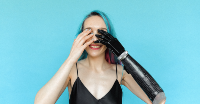 A woman with blue and pink hair and a prosthetic arm is smiling with her hands over her eye peek-a-boo style.