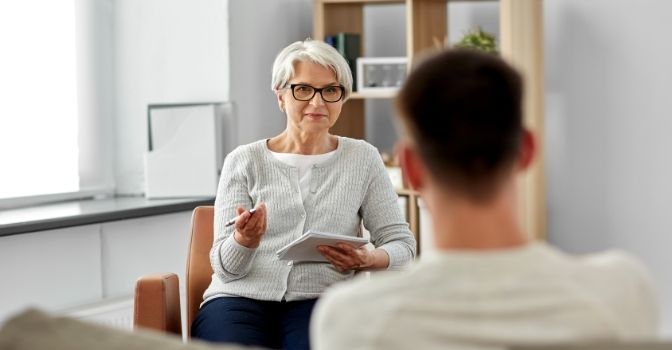 A psychologist sit in front of her client, smiling at them and holding a notebook and pen.