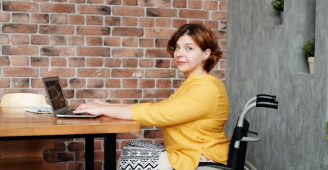 A lady in a wheelchair in a yellow top sits at a table with her laptop. Her head is turned and she is smiling at the camera.